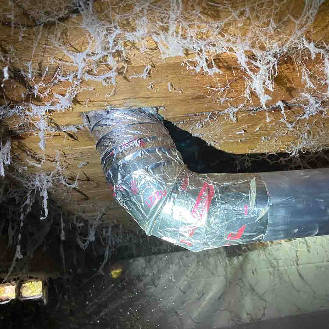 A silver metal dryer vent transition pipe in the crawlspace of a home. There is silver metal duct tape on the pipe where it has been repaired.