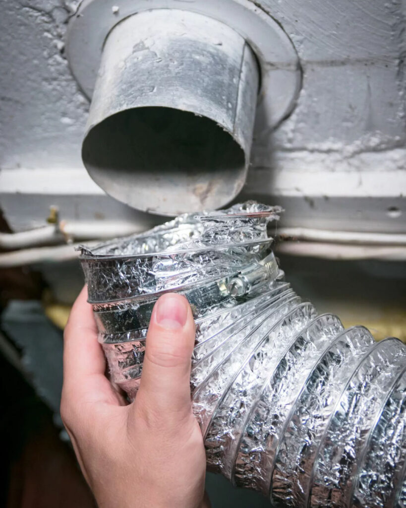 A technician installs a brand new silver metal dryer vent hose behind a dryer in a home. Environment is clean and white.