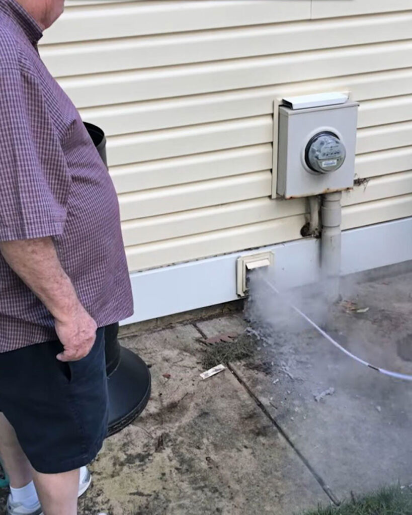A technician blows lint our of a dryer vent from outside a home. The lint creates a lint cloud at the opening as lint is forced out onto the sidewalk.