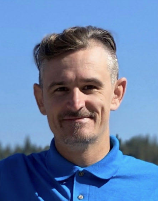 A medium shot of John Leslie the owner of Dryer Vent John. He is a youthful, Caucasian male with short goatee wearing a blue shirt. Behind him is a open, blue sky and green trees.